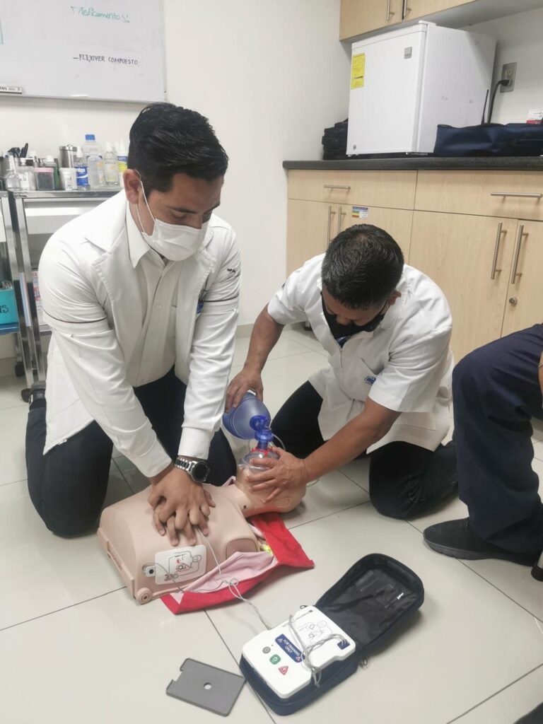 Dos profesionales de la salud demostrando RCP en un maniquí de entrenamiento médico, ambos con batas blancas, uno realizando compresiones torácicas y el otro usando una máscara de válvula de bolsa para proporcionar ventilación, en un entorno clínico con suministros y equipos médicos en el fondo, para la sección Capacitación de Personal y Brigadas de Salud Empresarial de México.